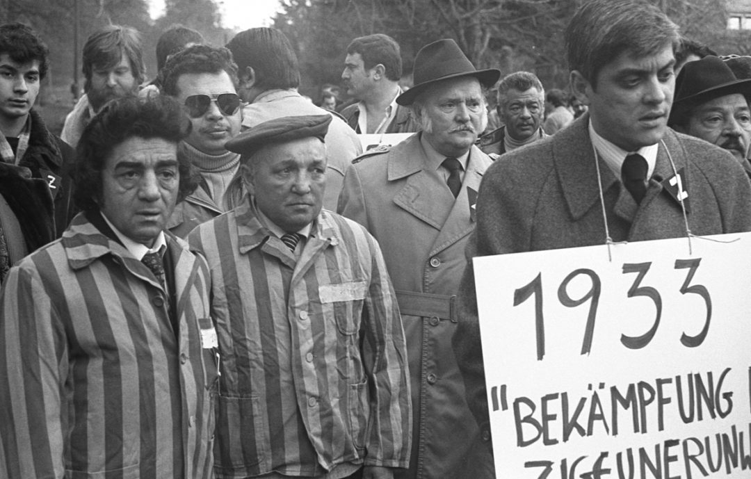 Historical black and white photo of protesting Sinti and Roma in front of the headquarters of the Federal Criminal Police Office in Wiesbaden. Romani Rose stands in the foreground on the right. He is wearing a sign around his neck. A black square with the letter "Z" is pinned to his lapel. To his left are two men who, as Holocaust survivors, have put on their striped prisoner's clothing from the concentration camp.
