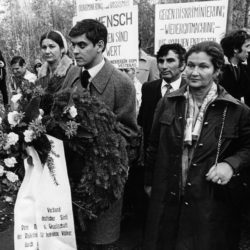 Romani Rose (links) und Simone Veil (rechts) bei einer Kranzniederlegung in Bergen-Belsen. Romani Rose trägt einen Kranz. Hinter den beiden sind weitere Teilnehmerinnen und Teilnehmer zu sehen, die Plakate mit Forderungen hochhalten.