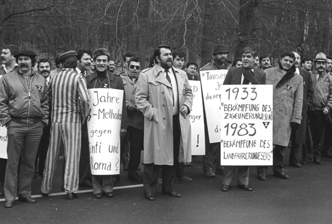 Historisches schwarz-weiß Foto von Sinti und Roma bei einem Protest vor der Zentrale des Bundeskriminalamtes in Wiesbaden. Mehrere Personen haben sich große Schilder mit Sprüchen umgehängt. Es nahmen auch Holocaust-Überlebende teil, die ihre gestreifte Häftlingskleidung aus den Konzentrationslagern trugen.