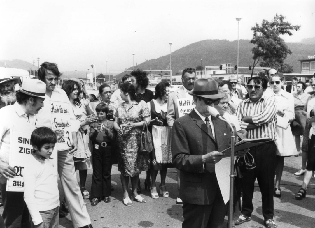 Vinzenz Rose stands in front of a microphone during a demonstration. Behind him in a semi-circle are other participants holding up placards.