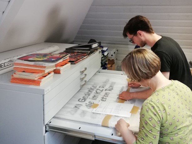 Eine Frau und ein Mann stehen vor einer ausgezogenen Schublade und blättern durch die Plakatsammlung des Dokumentationszentrums.