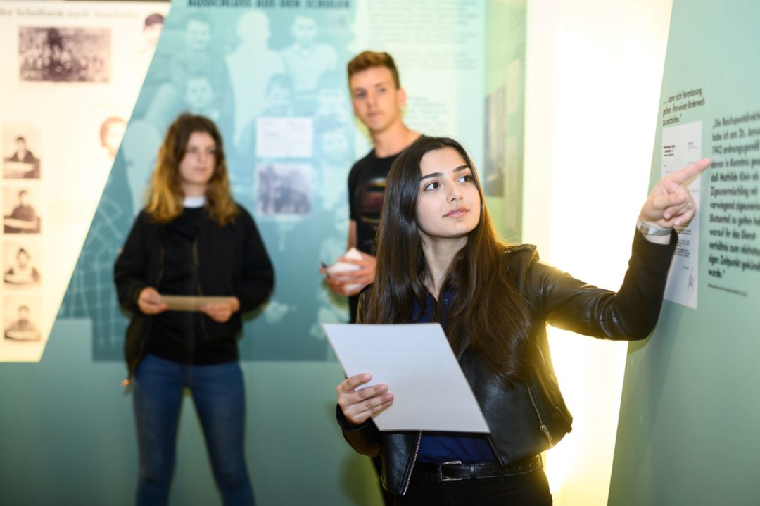 Young people during a workshop in the exhibition at the Documentation Center. In the foreground, a young woman points to a picture on an exhibition board during a presentation. In the background, two other young people can be seen listening.