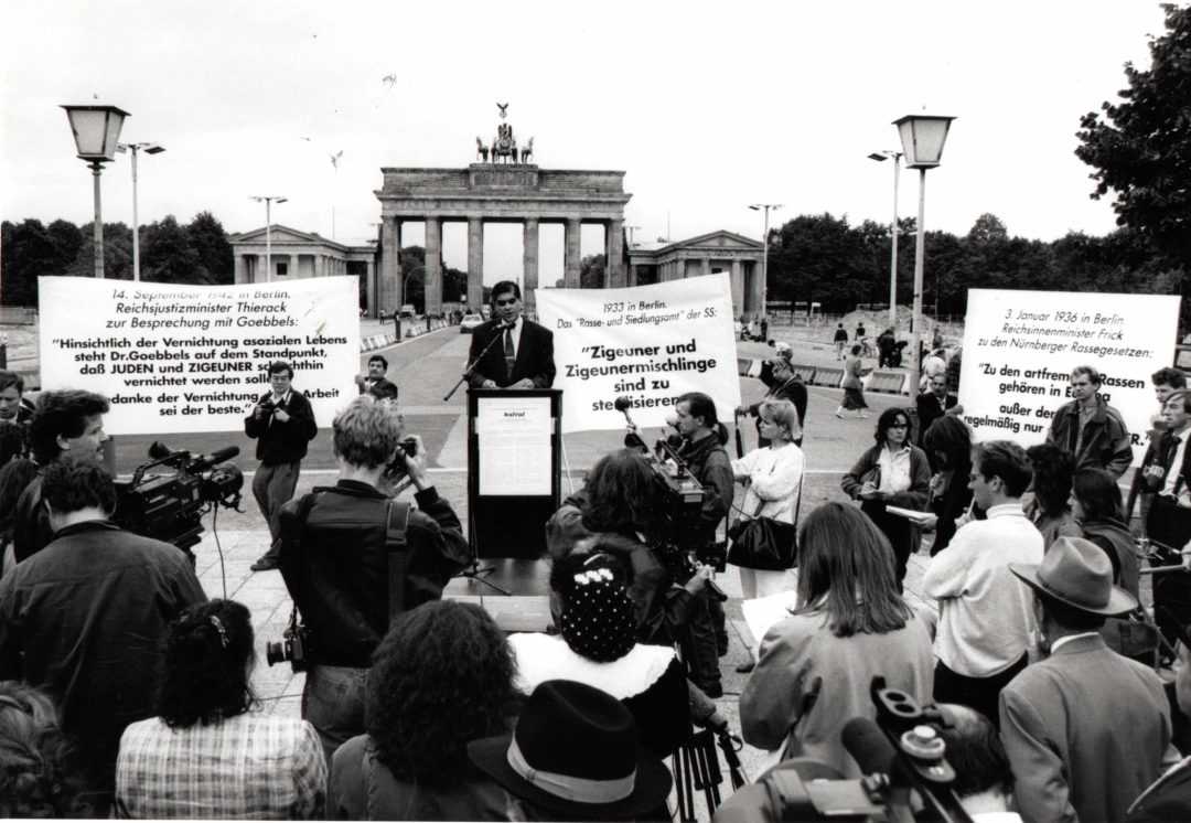 Historical black-and-white photo of a demonstration. In the foreground, participants of the demonstration stand with their backs to the camera, some holding up banners. In the center, Romani Rose stands at a raised lectern. The Brandenburg Gate in Berlin can be seen in the background.