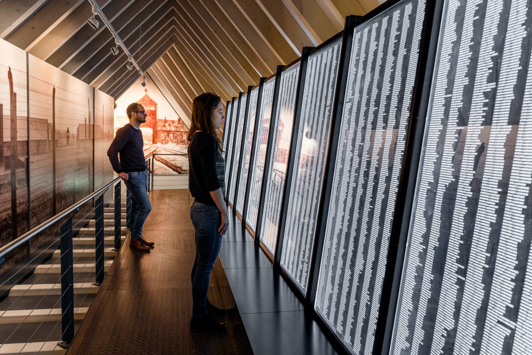Visitors in the permanent exhibition of the Documentation Center in Heidelberg. They stand in front of a long wall on which are written thousands of names of Sinti and Roma who were murdered in Auschwitz-Birkenau.