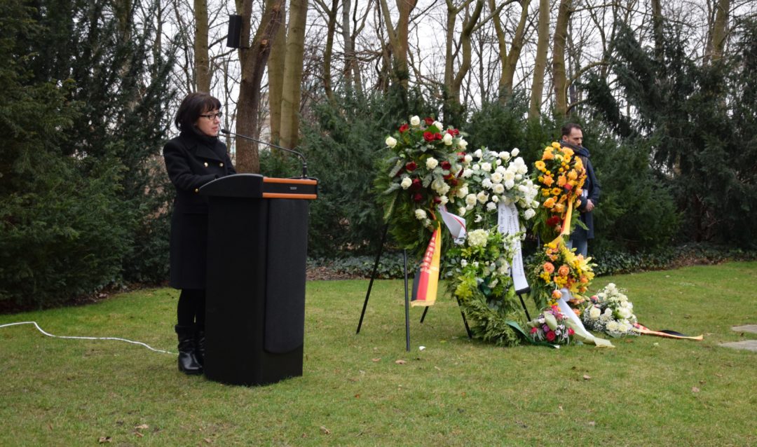 Petra Rosenberg an einem Rednerpult während ihrer Gedenkrede am Denkmal für die ermordeten Sinti und Roma Europas in Berlin. Neben ihr stehen mehrere große, aufgeständerte Gedenkkränze.
