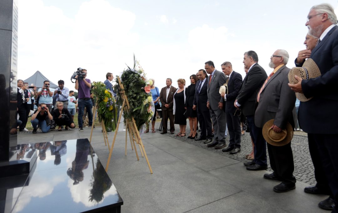 Eine Gruppe von Menschen steht vor dem Denkmal für die in Auschwitz-Birkenau ermordeten Sinti und Roma und verneigen sich. Vor dem Denkmal stehen zwei Gedenkkränze.