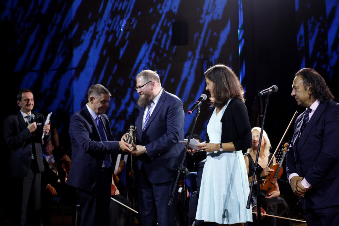Romani Rose hands over the statue of the Civil Rights Prize to Piotr Cywiński. Catharina Seegelken and Roman Kwiatkowski stand next to Piotr Cywiński. In the background are musicians from an orchestra