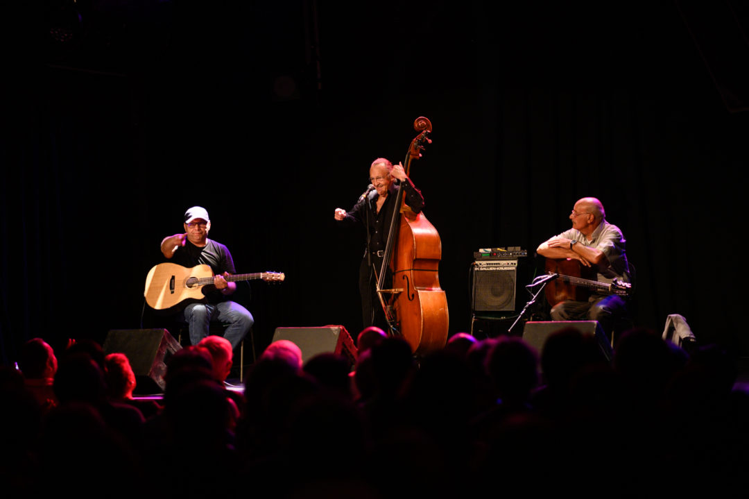 Three musicians on a stage. Two are sitting with guitars, between them stands the third on the double bass.