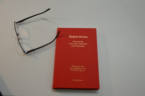 Foto des Buches "Djiparmissa. Übersetzung deutscher Gedichte in Romanes" von Reinhold Lagrene. Das Buch hat einen roten Einband, der Titel und der Autor sind vorne in goldener Farbe eingeprägt. Neben dem Buch liegt eine Lesebrille.