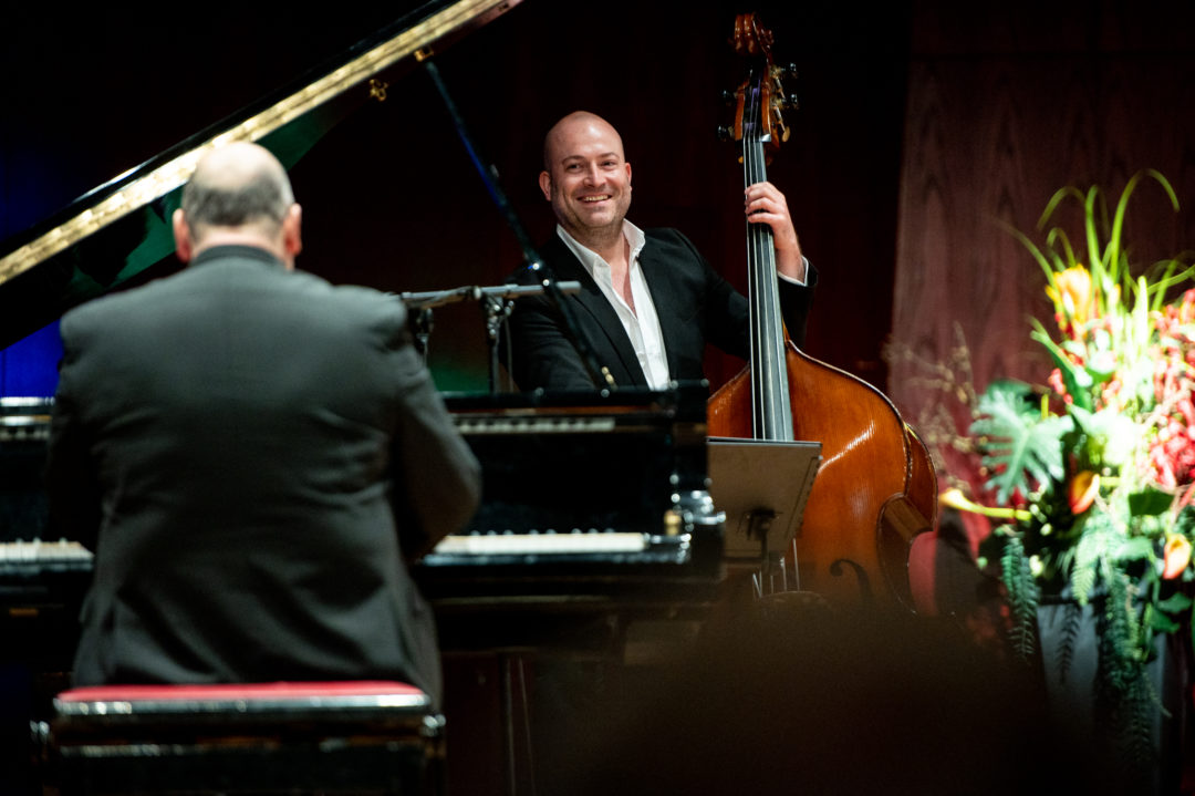 Zwei Männer auf einer Bühne. Im Vordergrund sitzt ein Mann mit dem Rücken zur Kamera an einem Flügel, dahinter steht ein Mann und spielt Kontrabass.