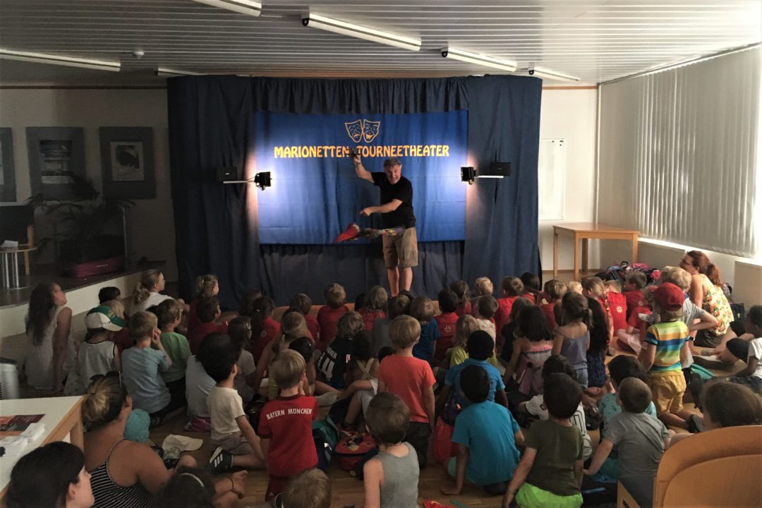 The puppeteer is standing on a stage. Many children are sitting on the floor in front of him.
