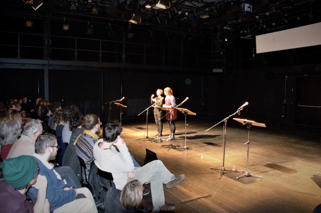 Zwei Frauen während der Lesung der "NSU-Monologe" auf einer Bühne. Vor ihnen stehen jeweils ein Mikrofon und ein Lesepult. Am linken Bildrand sitzt ein Teil des Publikums in Reihen.