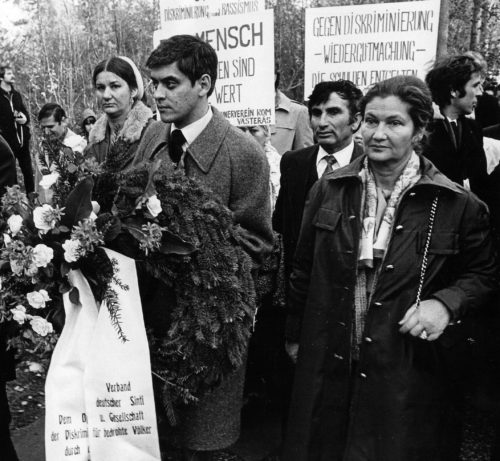 Romani Rose (links) und Simone Veil (rechts) bei einer Kranzniederlegung in Bergen-Belsen. Romani Rose trägt einen Kranz. Hinter den beiden sind weitere Teilnehmerinnen und Teilnehmer zu sehen, die Plakate mit Forderungen hochhalten.