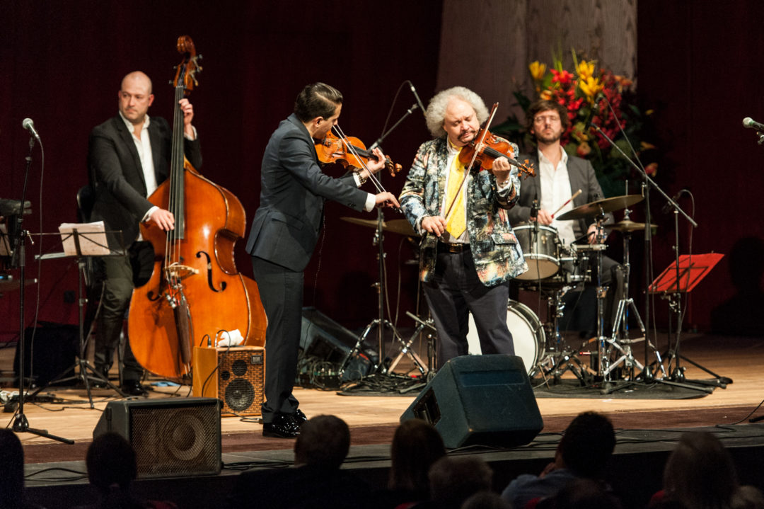 Vier Musiker auf einer beleuchteten Bühne. Im Vordergrund stehen zwei mit Violine, hinter ihnen links steht ein Mann am Kontrabass, rechts von ihnen sitzt ein Mann am Schlagzeug.
