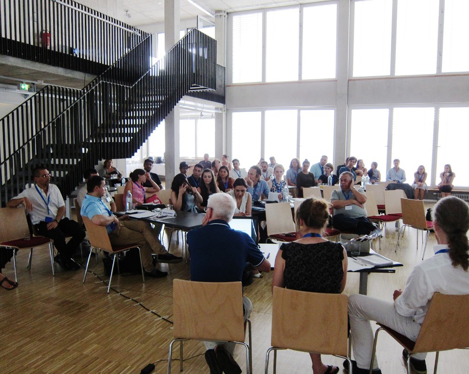 Blick in das Forum des Bildungsforum gegen Antiziganismus in Berlin. Darin sitzen viele Teilnehmer auf Stühlen zusammen und diskutieren.