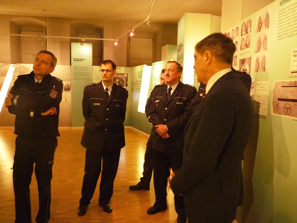Mehrere Polizisten in Uniform stehen um Romani Rose im Halbkreis in der Dauerausstellung. Romani Rose schildert Inhalte der Ausstellung.