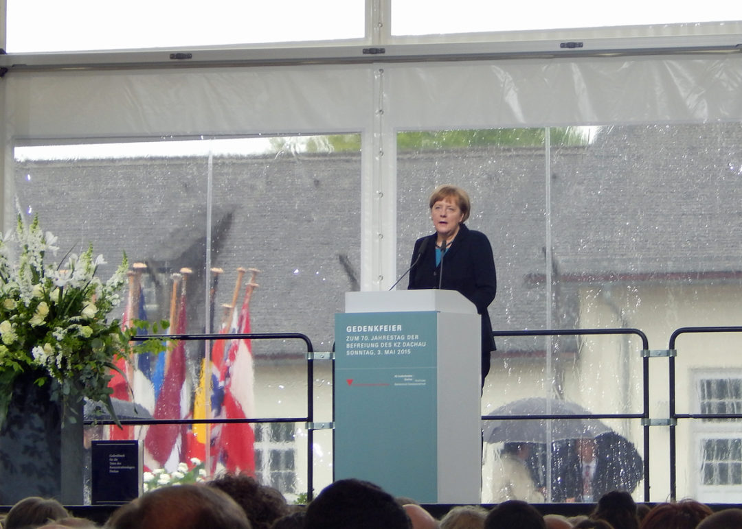Bundeskanzlerin Angela Merkel bei 
ihrer Ansprache. Sie steht an einem Rednerpult auf einer Bühne vor einem erhaltenen Gebäude in der KZ-Gedenkstätte Dachau.