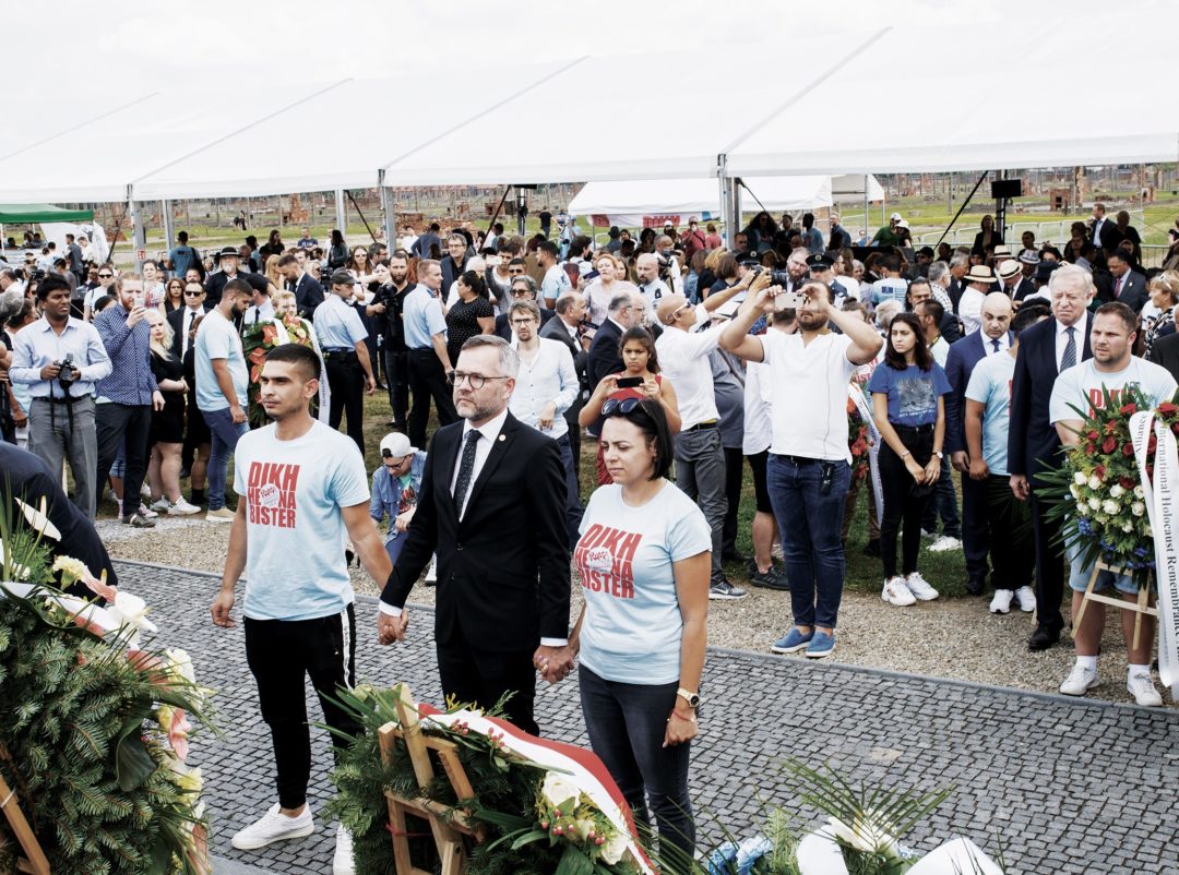 Drei Personen im Vordergrund stehen vor Trauerkränzen und halten sich an den Händen. Im Hintergrund steht eine Vielzahl von weiteren Teilnehmerinnen und Teilnehmern der Jugendgedenkfahrt.