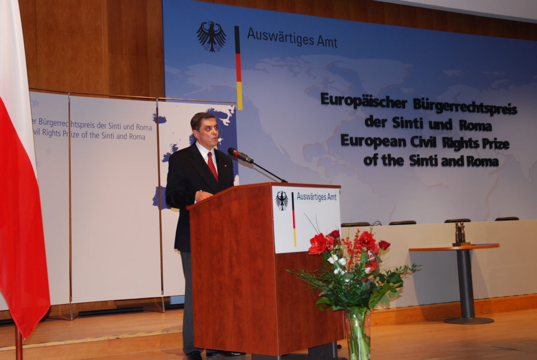 Romani Rose im Auswärtigen Amt während seiner Rede. Er steht an einem Rednerpult, links neben ihm stehen die europäische, die deutsche und die polnische Flagge. Rechts neben ihm ist der Titel der Veranstaltung auf einer großen Leinwand angebracht.