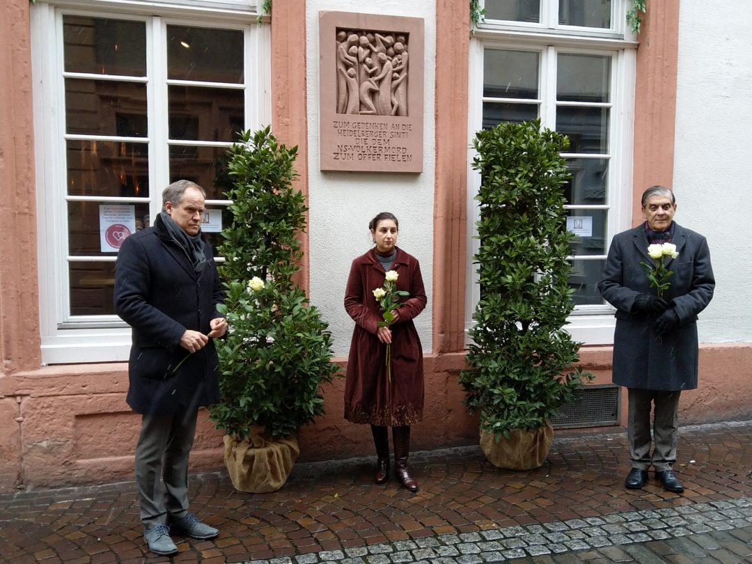 Oberbürgermeister Eckart Würzner, Dotschy Reinhardt und Romani Rose stehen vor der Gedenktafel für die Heidelberger verfolgten Sinti. Die Tafel aus Sandstein ist an einer Hauswand angebracht. Die drei halten jeweils eine Rose in den Händen.