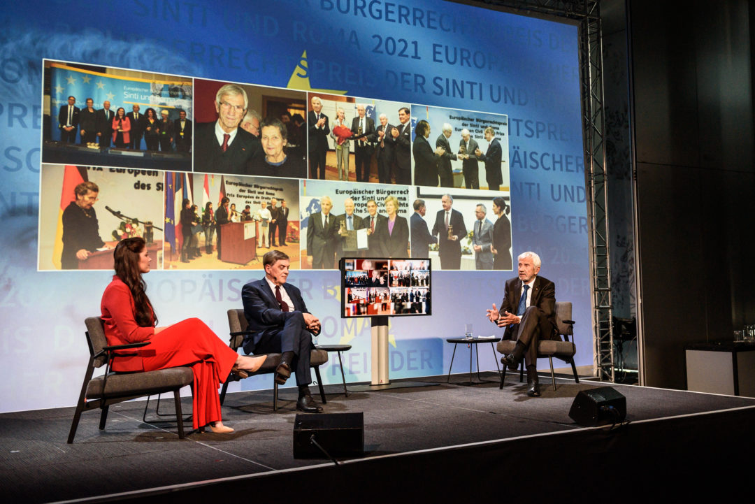 Angelina Kappler sits on the left in a red dress. In the middle sits Romani Rose, on the right Manfred Lautenschläger. The two are wearing suits. All three are in conversation. In the background is a collage of pictures from past award ceremonies.
