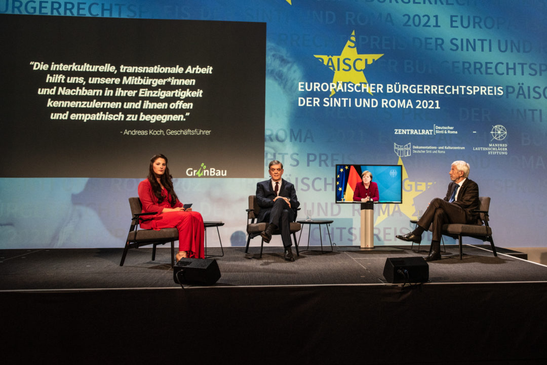 Presenter Angelina Kappler, Romani Rose and Manfred Lautenschläger sit together in conversation at a distance due to the corona pandemic. In the background, German Chancellor Angela Merkel is connected live via a screen.