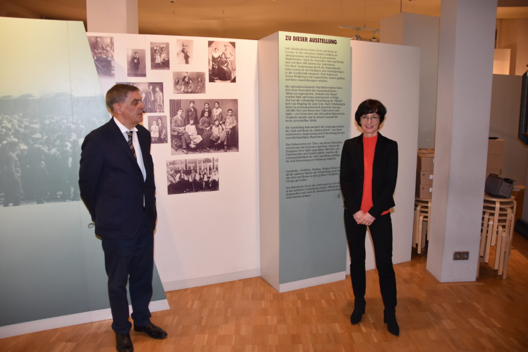 Romani Rose und Bürgermeisterin Stefanie Jansen während einer Führung durch die Dauerausstellung im Dokumentations- und Kulturzentrum. Sie stehen vor einer Ausstellungswand mit historischen Familienfotos von Sinti und Roma.