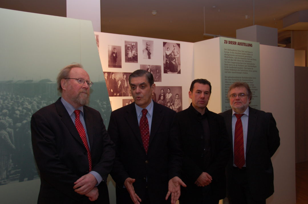 Von links: Wolfgang Thierse, Romani Rose und Silvio Peritore und eine unbekannte Personen stehen nebeneinander in der Dauerausstellung des Dokumentations- und Kulturzentrums. Im Hintergrund ist eine Ausstellungswand mit historischen Familienbildern von Sinti und Roma zu sehen.