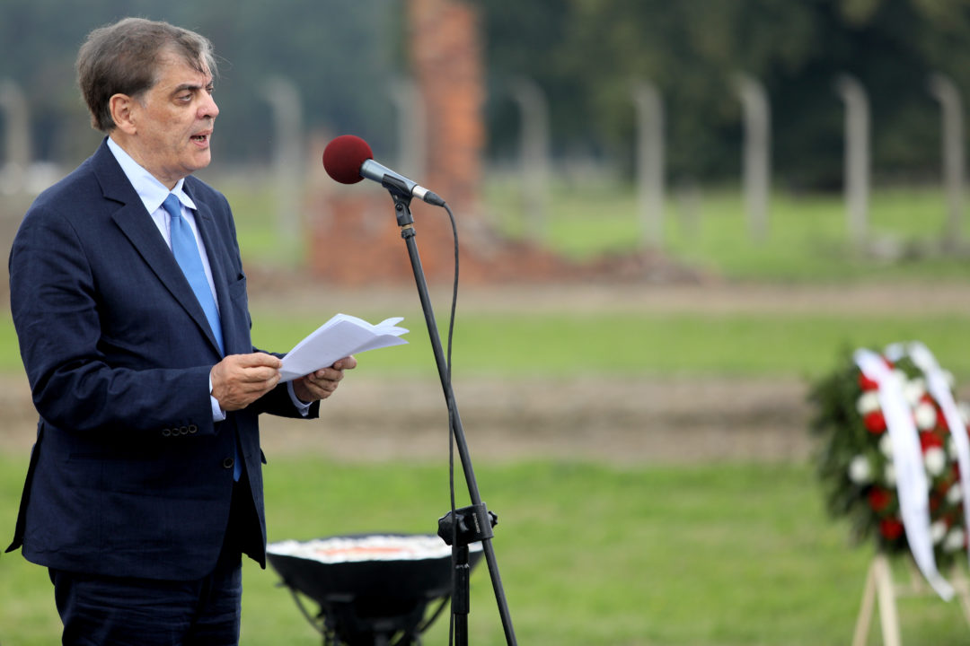 Romani Rose stands in front of a microphone and gives a speech on the grounds of the former Auschwitz-Birkenau death camp. A memorial wreath and remains of a fence can be seen in the background.