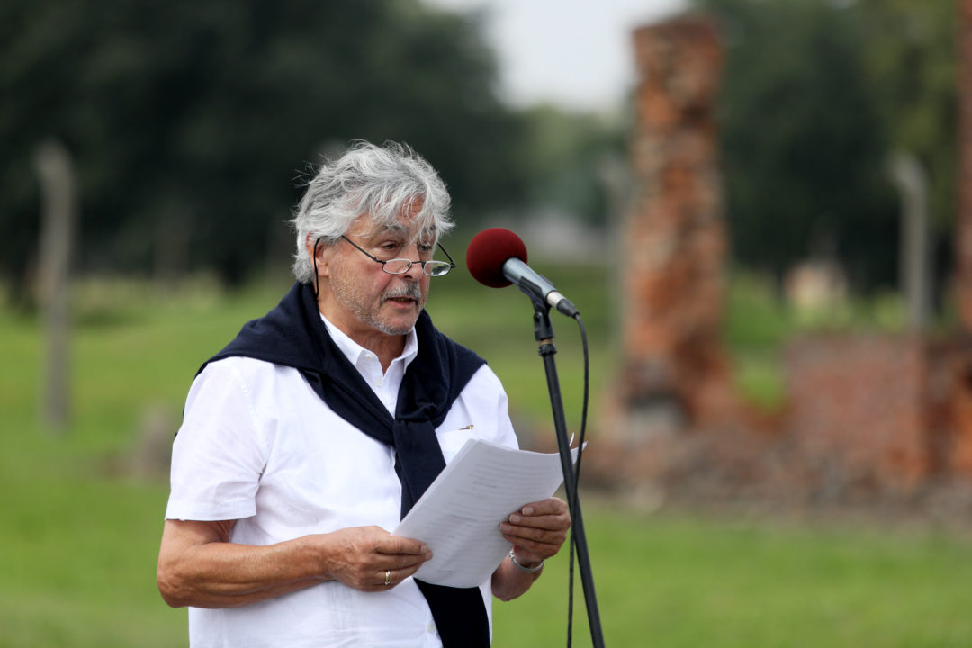 Holocaust survivor Christian Pfeil from Trier gave a moving speech at the memorial service at the former Auschwitz-Birkenau extermination camp. He is standing in front of a microphone. Remains of a camp barrack can be seen in the background.