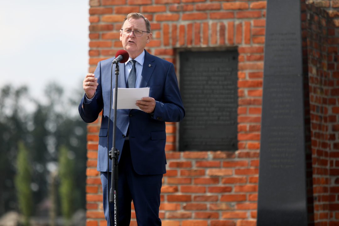 Bodo Ramelow stands in front of the memorial to the murdered Sinti and Roma in Auschwitz-Birkenau. He is speaking into a microphone. The memorial in the background is made of bricks. In front of the brick wall is a stele made of black stone.