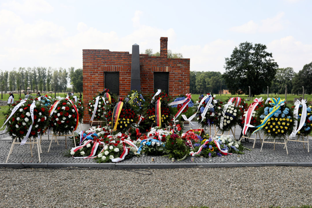In the foreground, dozens of wreaths made of flowers with commemorative bows can be seen. They lie and stand in front of the memorial to the murdered Sinti and Roma in Auschwitz-Birkenau. The memorial is made of bricks. Two black metal panels with text are embedded in it. Between the panels is a stele made of black stone.