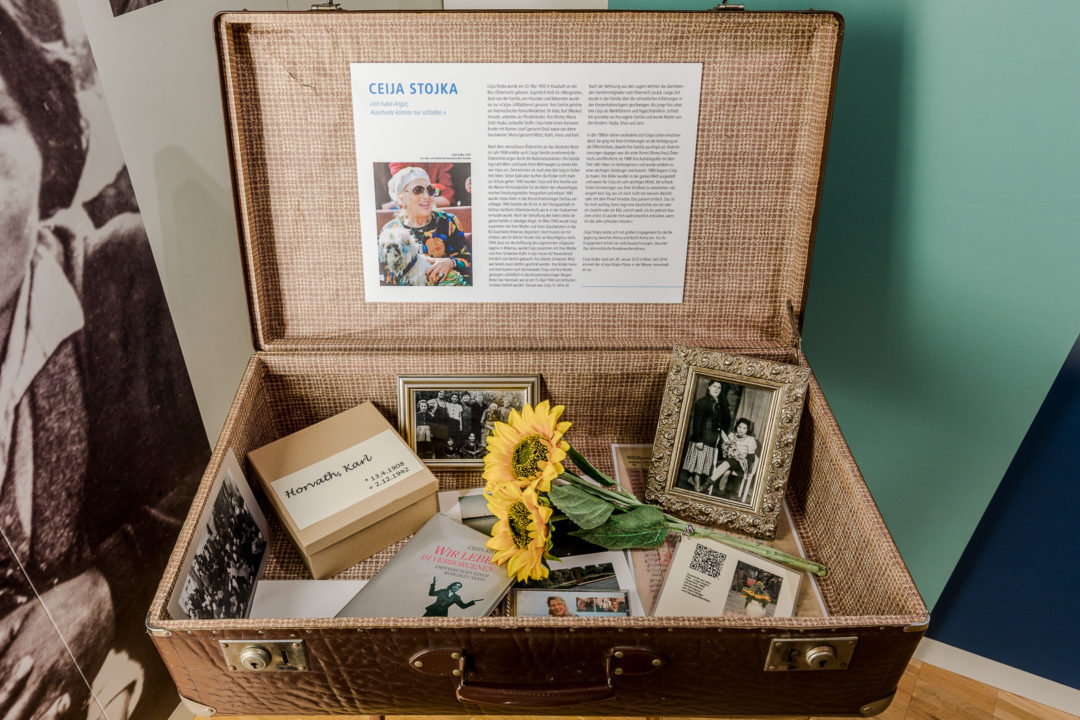 In einem alten Koffer sind zahlreiche Gegenstände, darunter Familienbilder, Bücher und ein Strauß aus Sonnenblumen. Im Deckel des Koffers sind ein Bild von Ceija Stojka und biografische Informationen angebracht.