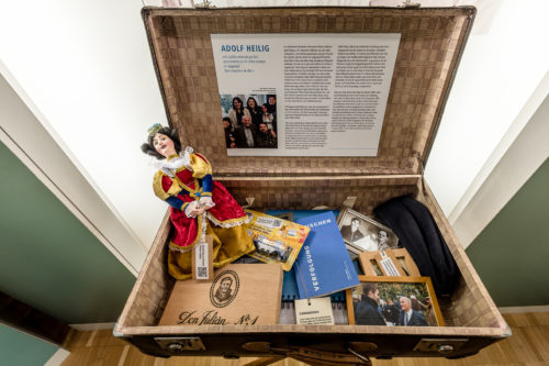 In einem alten Koffer sind zahlreiche Gegenstände, darunter Familienbilder, eine Zigarrenkiste, Bücher und eine farbenfrohe Handpuppe. Im Deckel des Koffers ist ein Bild von Adolf Heilig und biografische Informationen angebracht.