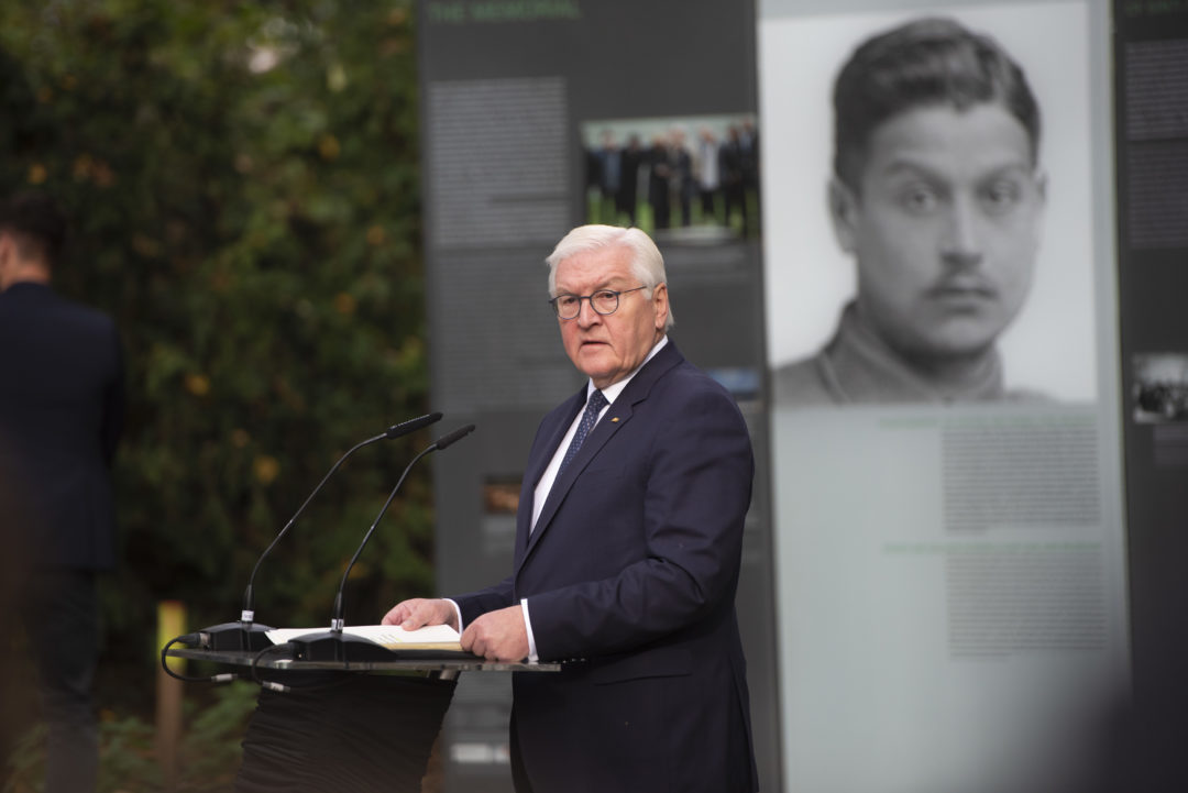 Bundespräsident Frank-Walter Steinmeier steht während seiner Gedenkrede an einem Rednerpult. Hinter ihm ist eine Freiluftausstellung mit Informationen zu individuellen Verfolgungsschicksalen von Sinti und Roma im Nationalsozialismus zu sehen.