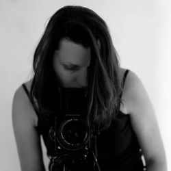 Valérie Leray blick in eine Fotokamera. Die Kamera ist zum Betrachter/zur Betrachterin hin ausgerichtet.
