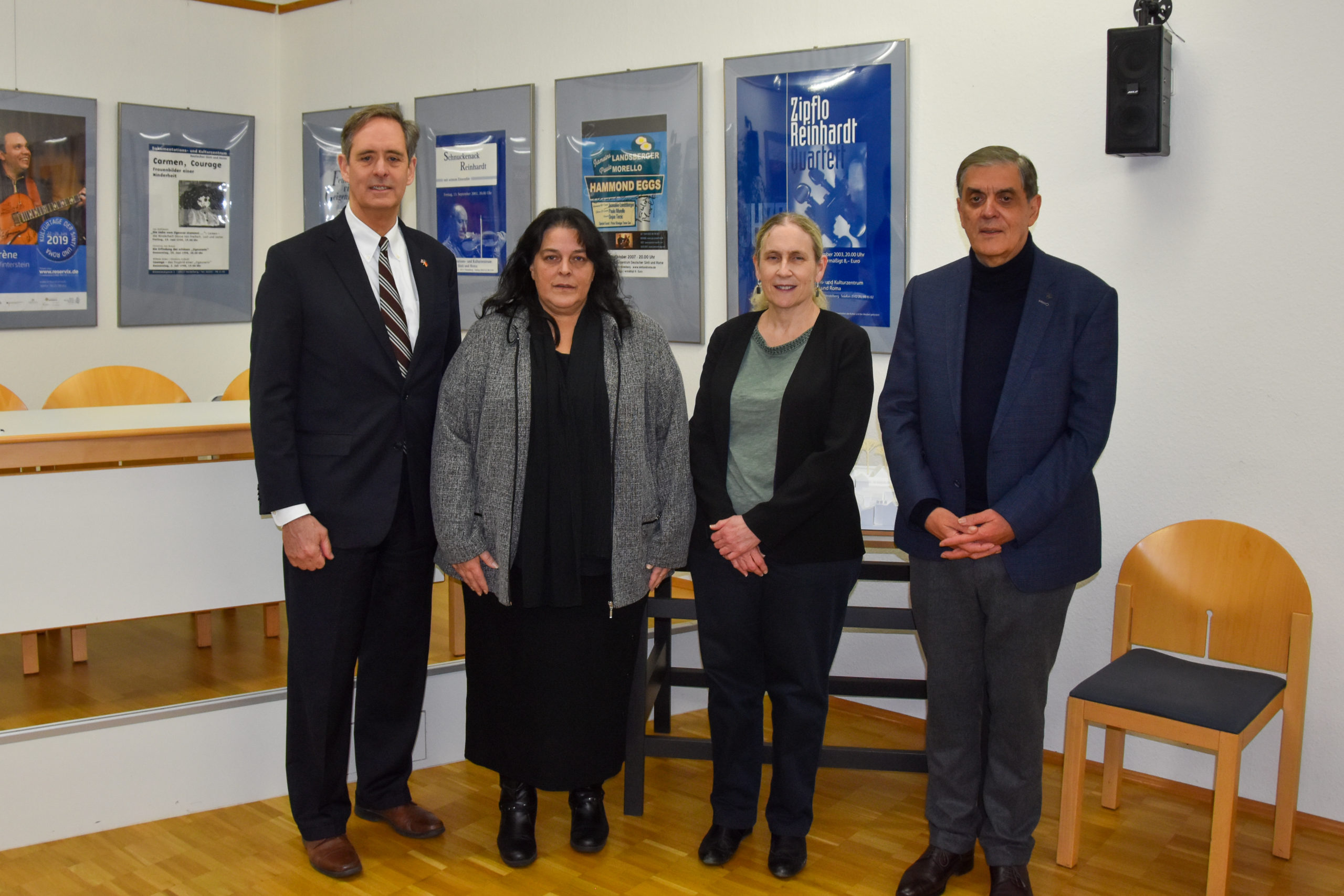 From left to right: Consul General Norman Thatcher Scharpf, Diana Bastian, Ambassador Ellen Germain and Romani Rose stand next to each other and look into the camera. In the background, a small stage with posters from the Documentation Center's event program can be seen.