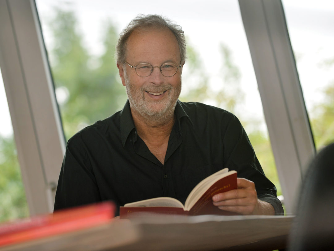Foto von Manfred Schmitz-Berg während einer Lesung. Er hält ein aufgeschlagenes Buch in seinen Händen und blickt lächelnd in die Kamera.
