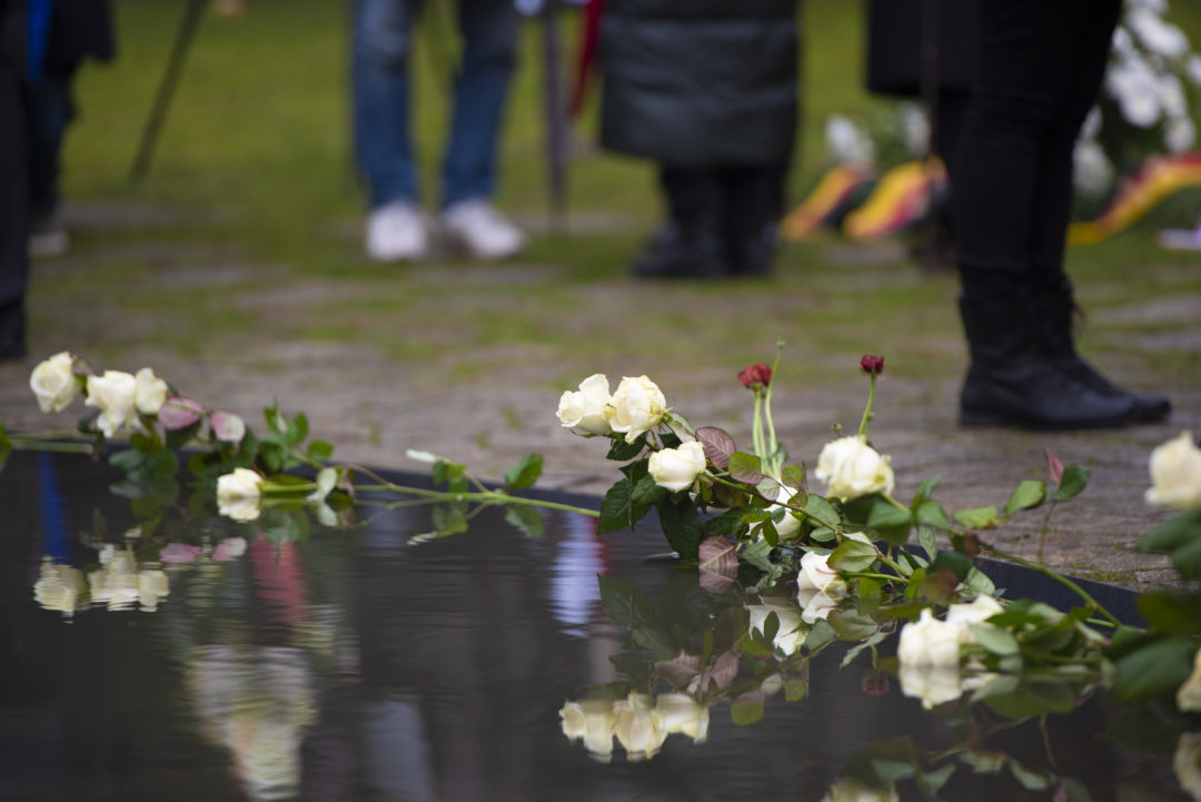 Eine Reihe von Rosen am Rand des schwarzen Wasserbeckens des Denkmal für die im Nationalsozialismus ermordeten Sinti und Roma Europas. Im Hintergrund sind verschwommen die Beine von Teilnehmerinnen und Teilnehmer der Gedenkveranstaltung zu erkennen.