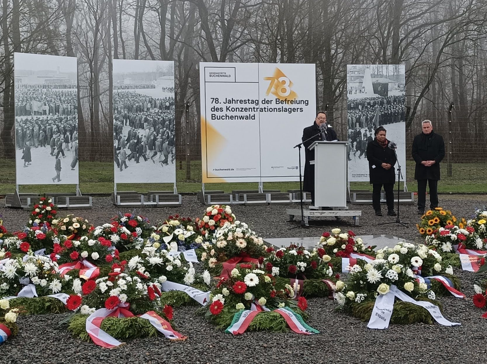 Jacques Delfeld stands at a lectern during his memorial speech. In front of him are many wreaths of flowers. Behind him are large-format posters with pictures of the liberation of Buchenwald concentration camp.