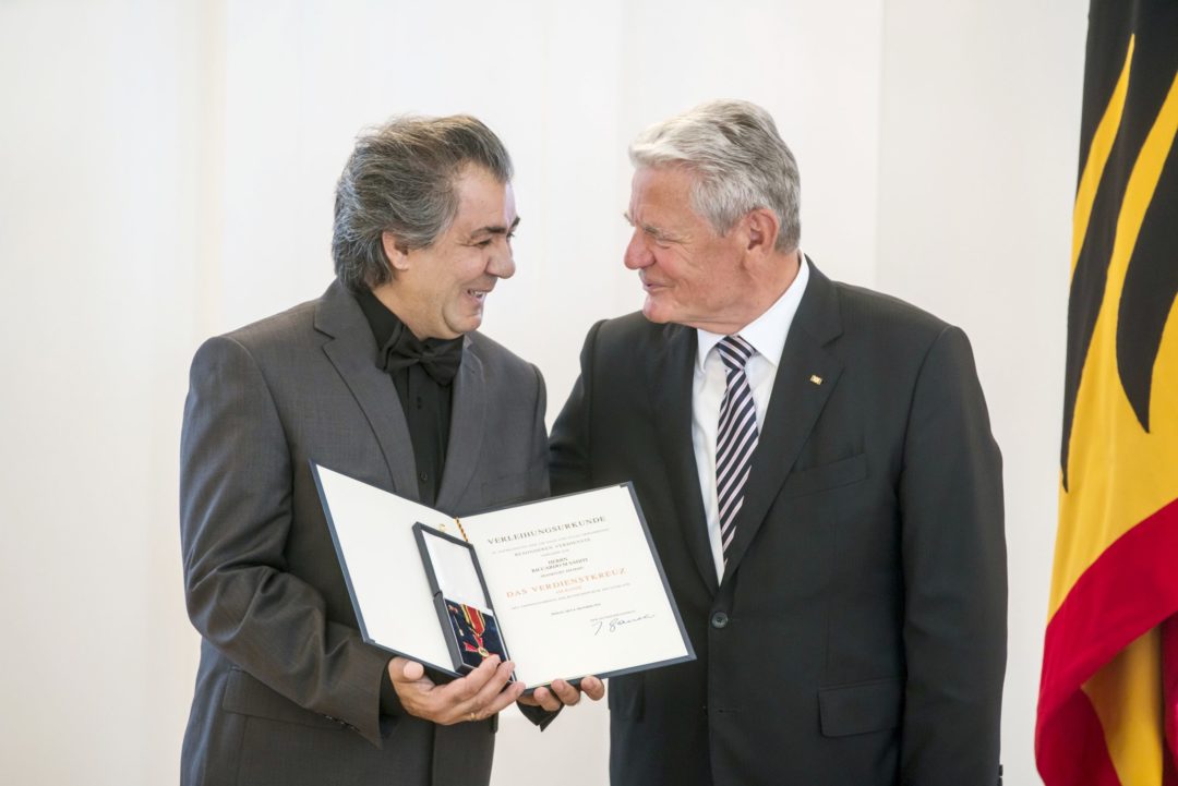 Riccardo M Sahiti hält das Bundesverdienstkreuz und eine Urkunde in seiner Hand. Rechts neben ihm steht der Bundespräsident Joachim Gauck. Die beiden schauen sich freundlich lächelnd an.
