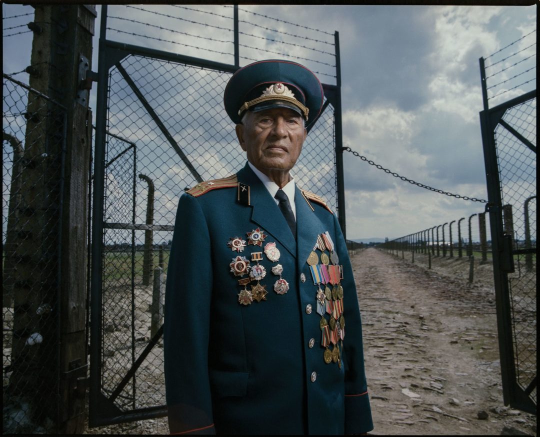 Ivan Bilashchenko stands in front of a gate in Auschwitz-Birkenau in his Red Army uniform with many medals.