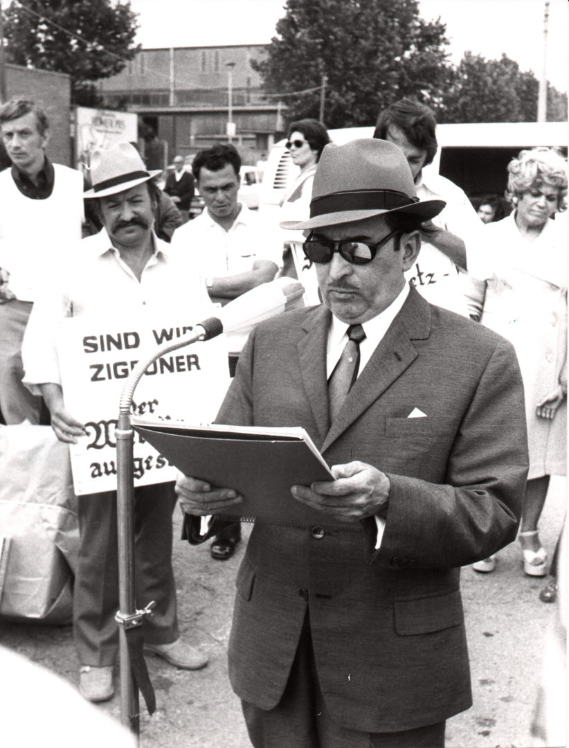 Das Foto zeigt Vinzenz Rose bei seiner Ansprache auf dem Alten Messplatz in Heidelberg. Er trägt einen Anzug, Hut und Sonnenbrille. Er steht vor einem Mikrofon. Hinter ihm stehen Demonstierende mit Schildern in den Händen.