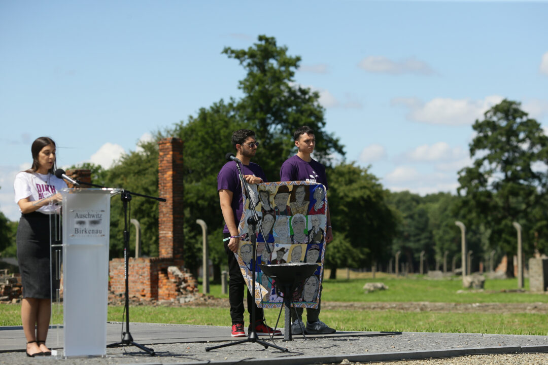 Georgina Laboda stands at a lectern during her memorial speech. Next to her, two other participants of the youth memorial event "Dikh He Na Bister" can be seen holding up a banner with portraits of Holocaust survivors.