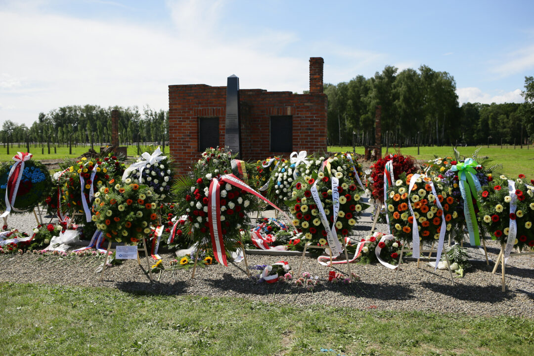 In the foreground, dozens of wreaths made of flowers with commemorative bows can be seen. They lie and stand in front of the memorial to the murdered Sinti and Roma in Auschwitz-Birkenau. The memorial is made of bricks. Two black metal plaques with text are embedded in it. Between the panels is a stele made of black stone.