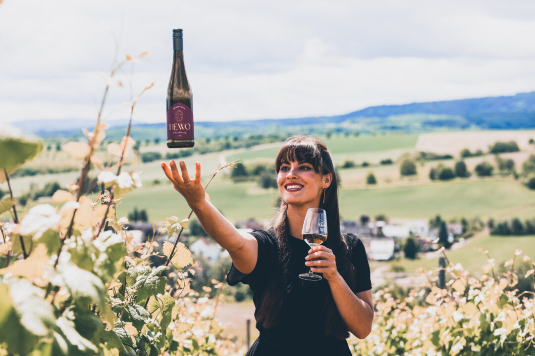 Angelina Kappler steht in einen Weinberg. In der einen Hand hält sie ein Weinglas, mit der anderen Hand wirft sie eine Weinflasche hoch, sodass es so aussieht, als würde sie die Flasche mit ihrer Hand schweben lassen.