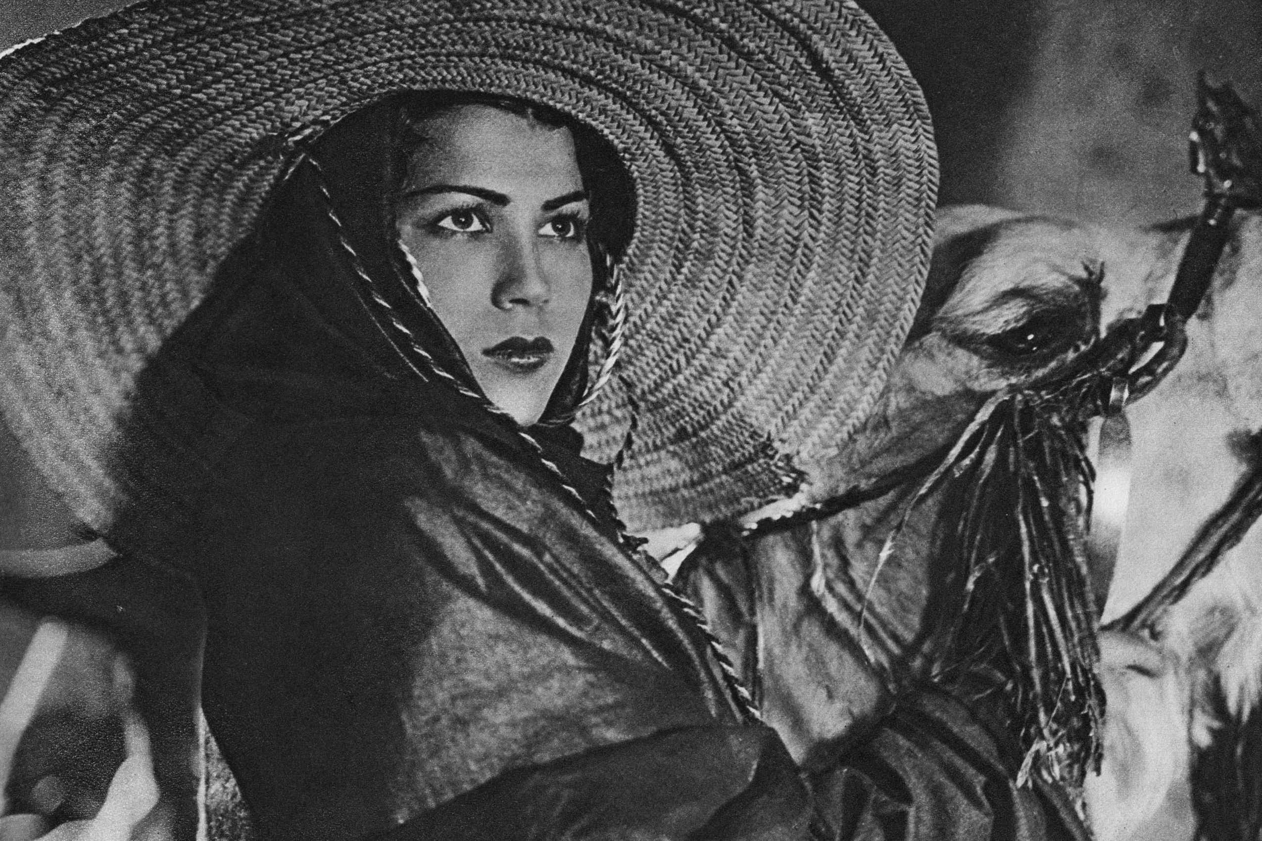 Ausschnitt aus einem alten schwarz-weiß Film mit der Schauspielerin Tela Tchaï. Sie trägt einen großen Strohhut. Sie hält ein Kamel am Zügel.