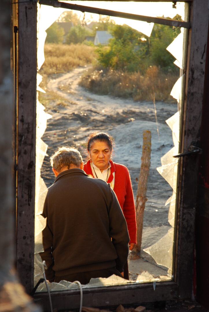 Ágnes Daróczi vor dem zerstörten Haus einer Roma-Familie in Ungarn. Sie schaut durch eine zerbrochene Fensterscheibe in das haus hinein. Vor dem Fenster steht ein Bewohner des Hauses mit dem Rücken zur Kamera gewandt.