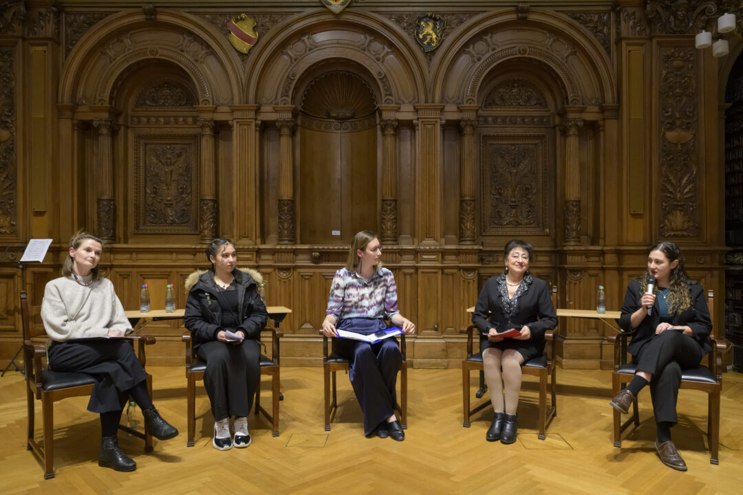 Fünf Frauen sitzen während einer Podiumsdiskussion nebeneinander. Eine Frau ganz rechts hält ein Mikro und spricht, die anderen blicken zu ihr. Das Bild wurde im festlichen Großen Rathaussaal Heidelberg vor einer holzgetäfelten Wand aufgenommen.