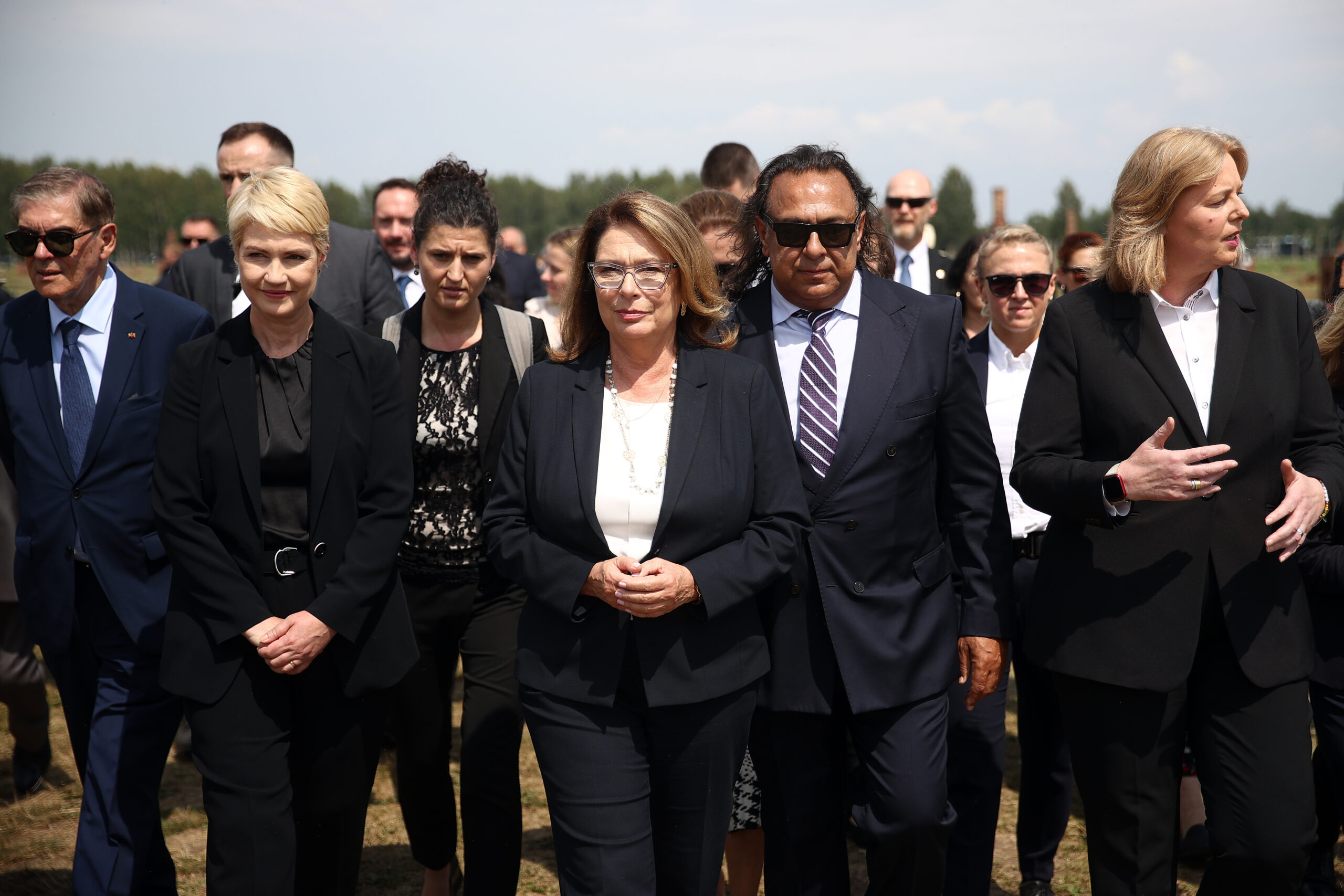 A group of people walk side by side along a stony dirt track in the former camp section B II e of the Auschwitz-Birkenau extermination camp. The gate of the section can be seen in the background, through which the group had to pass on their way to the memorial to the murdered Sinti and Roma. All the people are wearing black clothing.